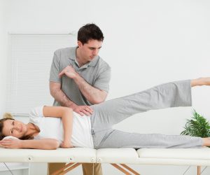 Doctor pressing his elbow on her hip while woman raising her leg in a room