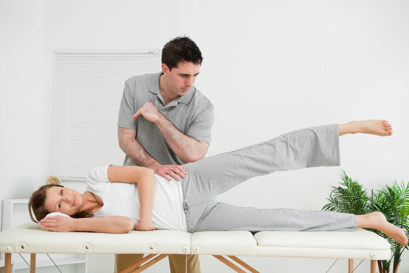 Doctor pressing his elbow on her hip while woman raising her leg in a room